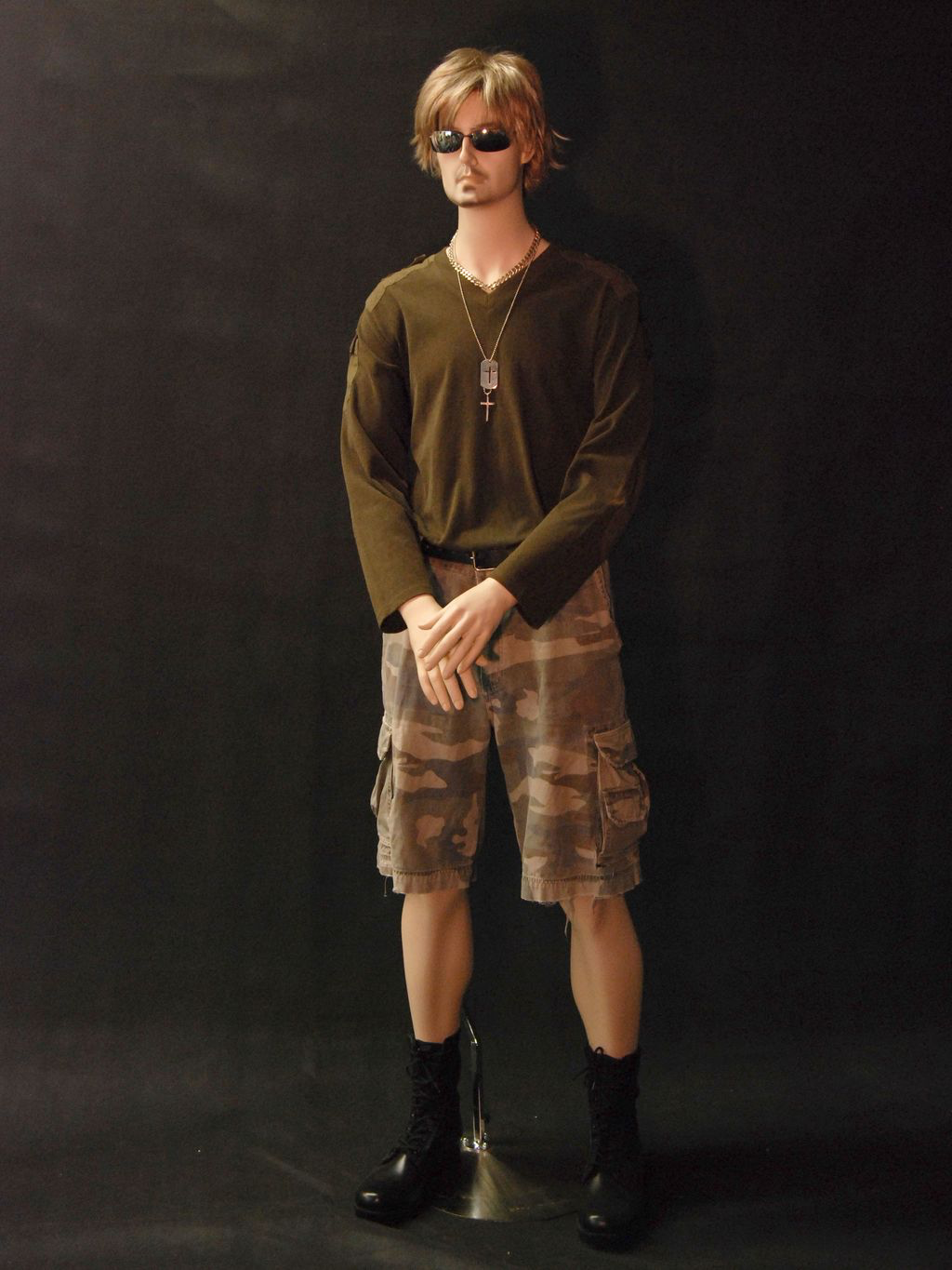 Person wearing a green long-sleeve shirt, camouflage shorts, and black boots against a dark background